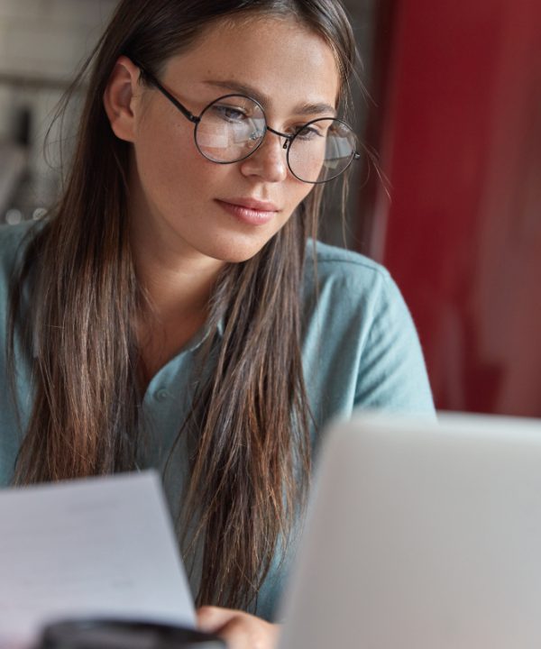 close-up-shot-freckled-woman-big-round-spectacles-examines-paper-work-checks-business-project-laptop-computer-drinks-aromatic-coffee-poses-indoor-reads-recommendation-fro